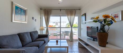 A living room with a grey sofa, a white coffee table, a TV on a stand, and a vase with flowers.