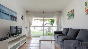 Un salon moderne doté d’un téléviseur à écran plat, d’une table basse blanche, d’un canapé gris et d’une porte-fenêtre coulissante donnant sur une terrasse aménagée avec du mobilier d’extérieur.