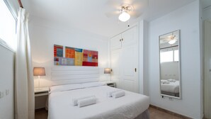 Une chambre d’hôtel avec un grand lit, deux tables de chevet avec des lampes, un ventilateur de plafond, un miroir et une œuvre d’art colorée accrochée au mur.