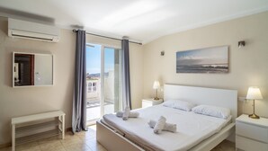 A hotel room with a large bed, a bedside table with a lamp, a bench, a sliding glass door to the balcony, and a painting on the wall.