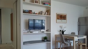 Ett modernt vardagsrum med en platt-TV, en matplats med bord och stolar samt ett köksutrymme med kylskåp.