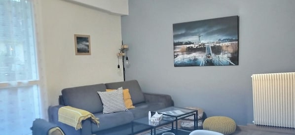 A living room with a grey sofa, a coffee table, a radiator, and a painting on the wall. - Nant'Sud Loire -NANTES -57m² new-2 bedrooms + sofa -10mn Airport & 12mn center (Nantes)