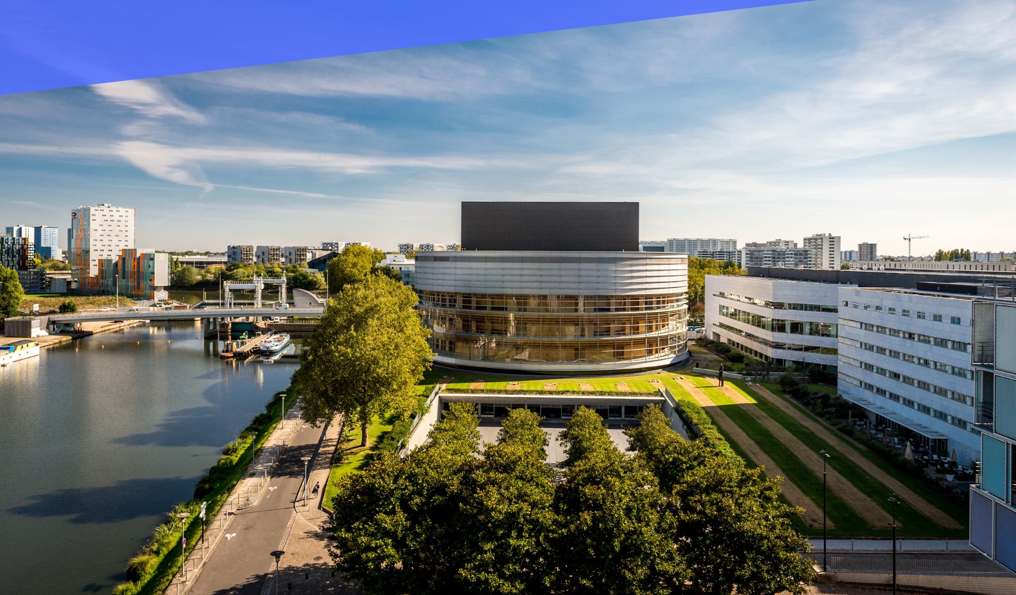A modern building with a curved facade, a river in the foreground, and a bridge in the background.