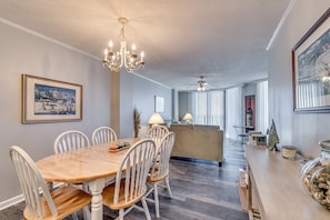 A dining area with a wooden table and white chairs, a chandelier, and a painting on the wall.