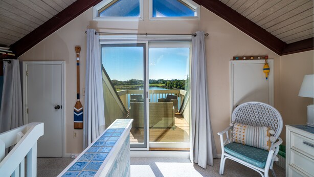 Habitación con un amplio ventanal corredero que da a un balcón con vistas a una piscina y un paisaje. Se observa una silla de mimbre con cojines y una mesita con mantel de estampado azul.