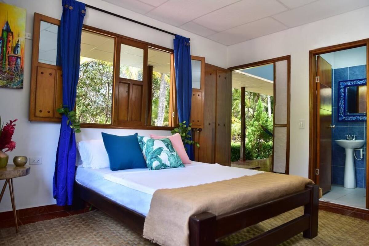 A bedroom with a bed, curtains, a nightstand, a painting, and a view of the outdoors.