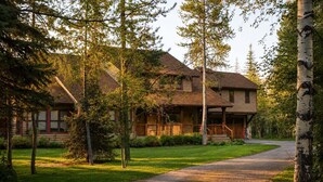 A two-story log cabin with a porch, surrounded by trees and a gravel driveway. - RMR: Mallory House in The Aspens/Wilson,WY (Wilson)