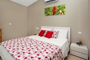 Une chambre à coucher avec un lit, du linge de lit blanc, des oreillers rouges, une table de chevet avec une plante et un climatiseur fixé au mur.