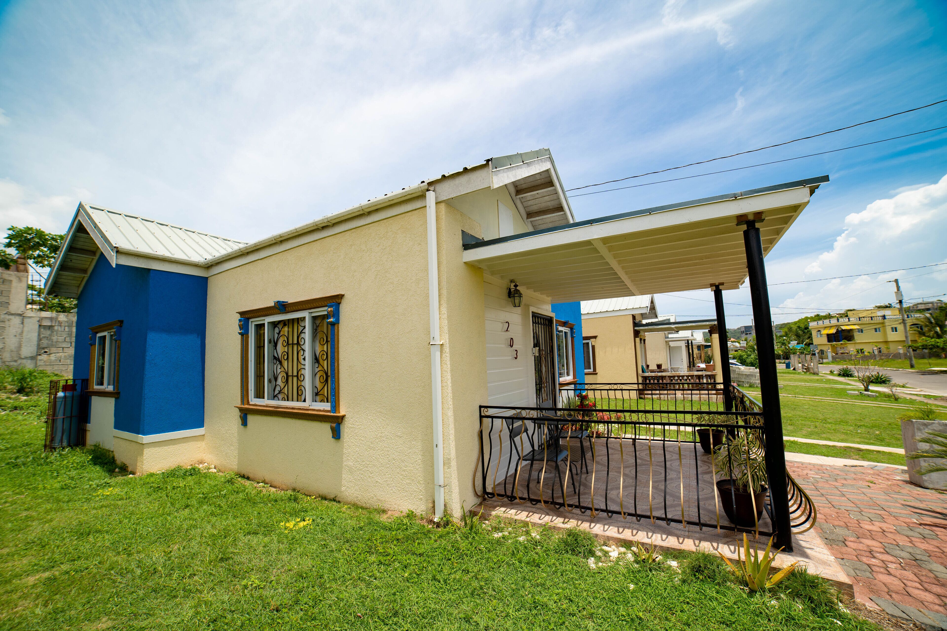 Une maison de plain-pied à l’extérieur bleu et beige, avec un porche couvert doté d’une balustrade noire et un petit jardin devant.