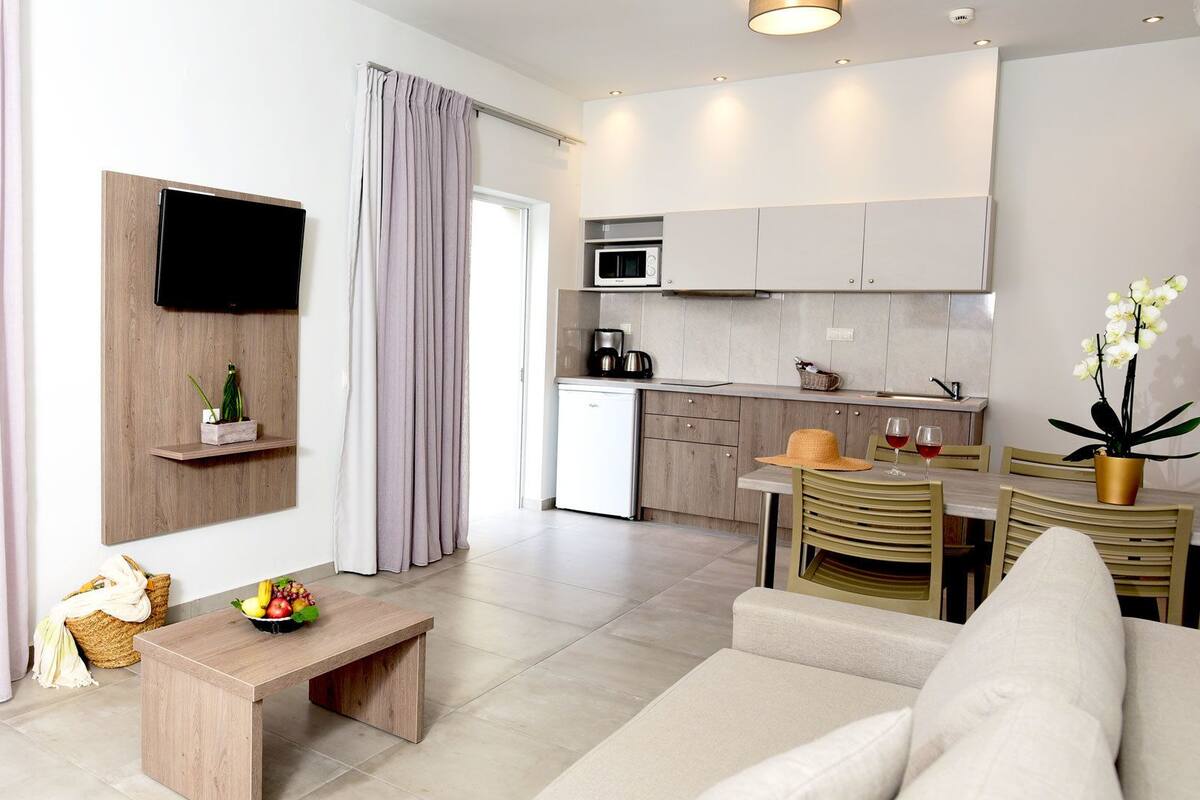 Uma sala moderna com sofá, mesa de centro de madeira com frutas, televisão fixada em um painel de madeira, e uma área de cozinha com micro-ondas e armários.