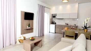 A modern living room with a sofa, a wooden coffee table with fruit, a TV mounted on a wooden panel, and a kitchen area with a microwave and cabinets.