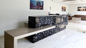 A reception desk with a marble top and a wooden bench in a modern lobby with a white floor and artwork on the walls. - Blue Horizon Apartments (Mylopotamos)