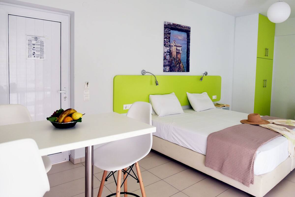 Quarto de hotel com uma cama, uma escrivaninha e uma cadeira. Há uma fruteira na escrivaninha e um quadro na parede.