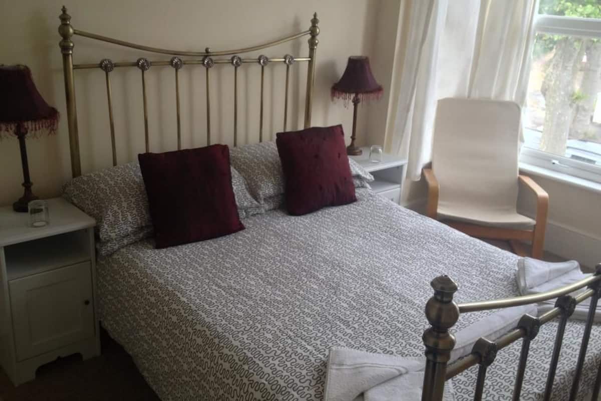 Una cama bien tendida con una colcha estampada, dos almohadas, una lámpara de noche y una silla cerca de una ventana.