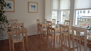 Un espace repas avec des tables et des chaises en bois, des tableaux encadrés au mur et de grandes fenêtres donnant sur l’extérieur.