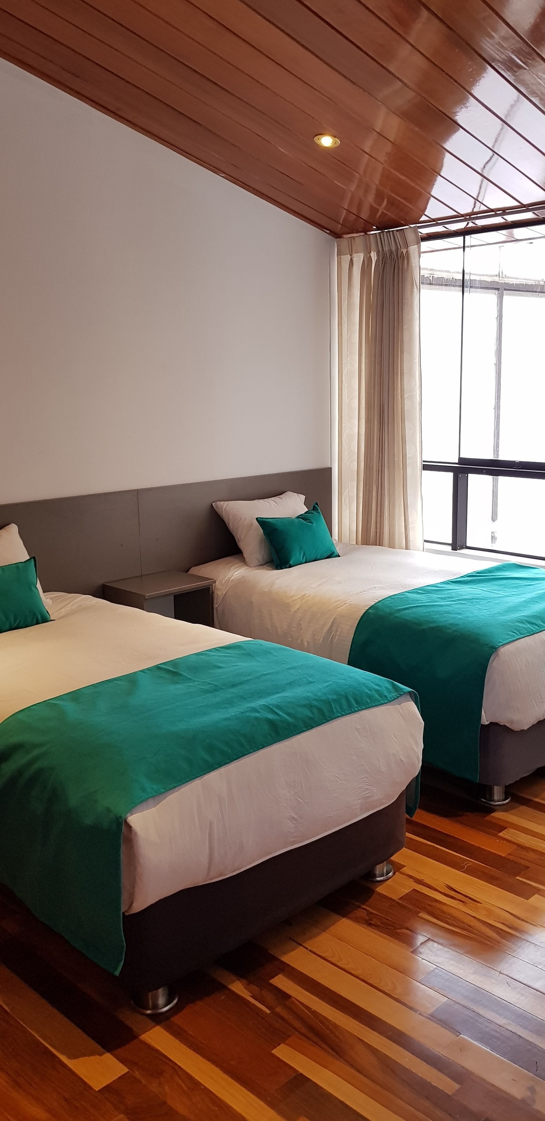 A hotel room with two beds, wooden flooring, a large window with curtains, and a wooden ceiling.