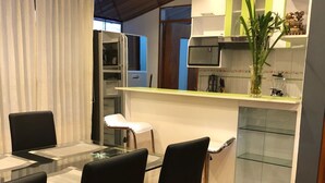 Un comedor moderno con una mesa de vidrio y sillas negras, una cocina con microondas y una planta, y suelo de madera.