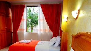 A hotel room with two beds, red curtains, a window with a view of greenery, and a wooden headboard. - Pampa & Selva Hotel (Rurrenabaque)