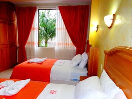 Habitación de hotel con dos camas, cortinas rojas, una ventana con vista a zonas verdes y un cabecero de madera.