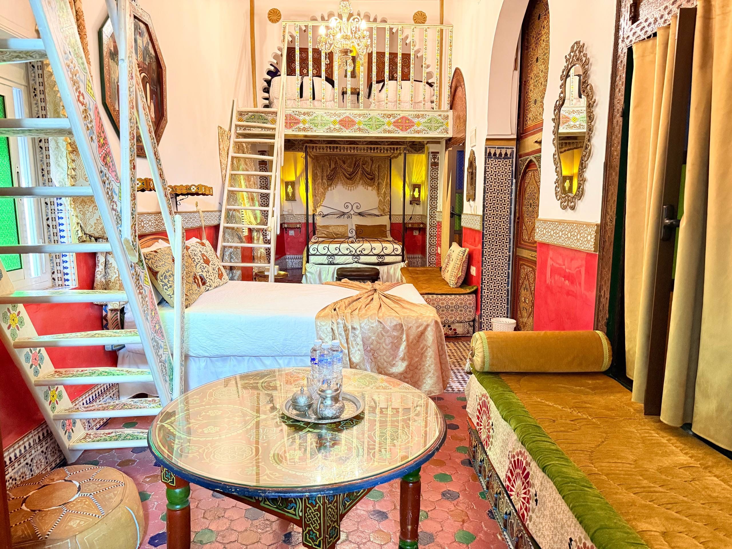 A room with a bed, a table with a decorative glass top, a sofa, and a staircase with patterned railings.
