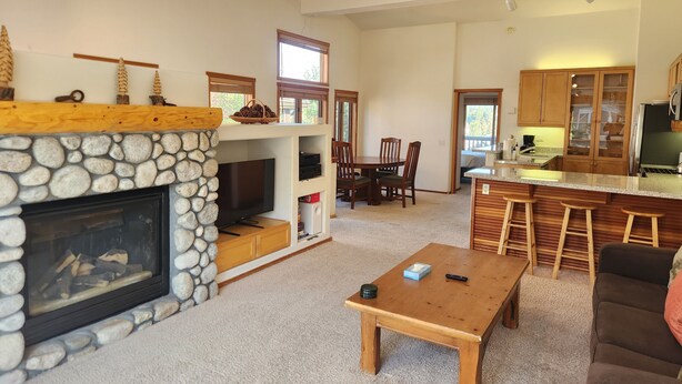 A living room with a stone fireplace, a dining area with wooden chairs, a kitchen with wooden cabinets, and a bar area with stools.
