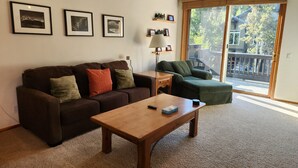 A living room with a brown sofa, a wooden coffee table, and a green armchair. There are framed pictures on the wall and a lamp on a side table.
