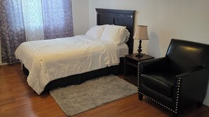 Un dormitorio con cama, una silla, una mesita de noche, una lámpara y una ventana con cortinas.
