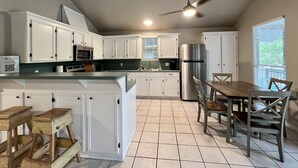 Eine Küche mit weißen Schränken, einem Edelstahlkühlschrank, einem Deckenventilator und einem Essbereich mit Holztischen und einem Tisch.