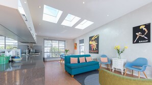 A modern living room with a blue sofa, green armchair, and a white side table with a vase of flowers. There are two paintings on the wall and a kitchen area in the background. - Designer house in quiet street on city doorstep (Darlinghurst)
