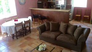 Un salón con un sofá marrón, una mesa de centro de madera con flores, una mesa de comedor con sillas, una zona de cocina con electrodomésticos y un reloj en la pared.