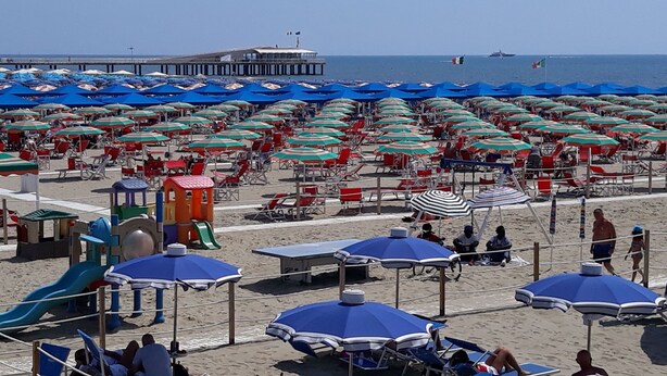Ein Strand mit vielen Sonnenschirmen und Liegestühlen, einem Spielplatz und einem Pier im Hintergrund.