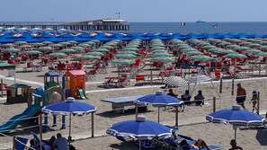 A beach with numerous umbrellas and chairs, a playground, and a pier in the background.