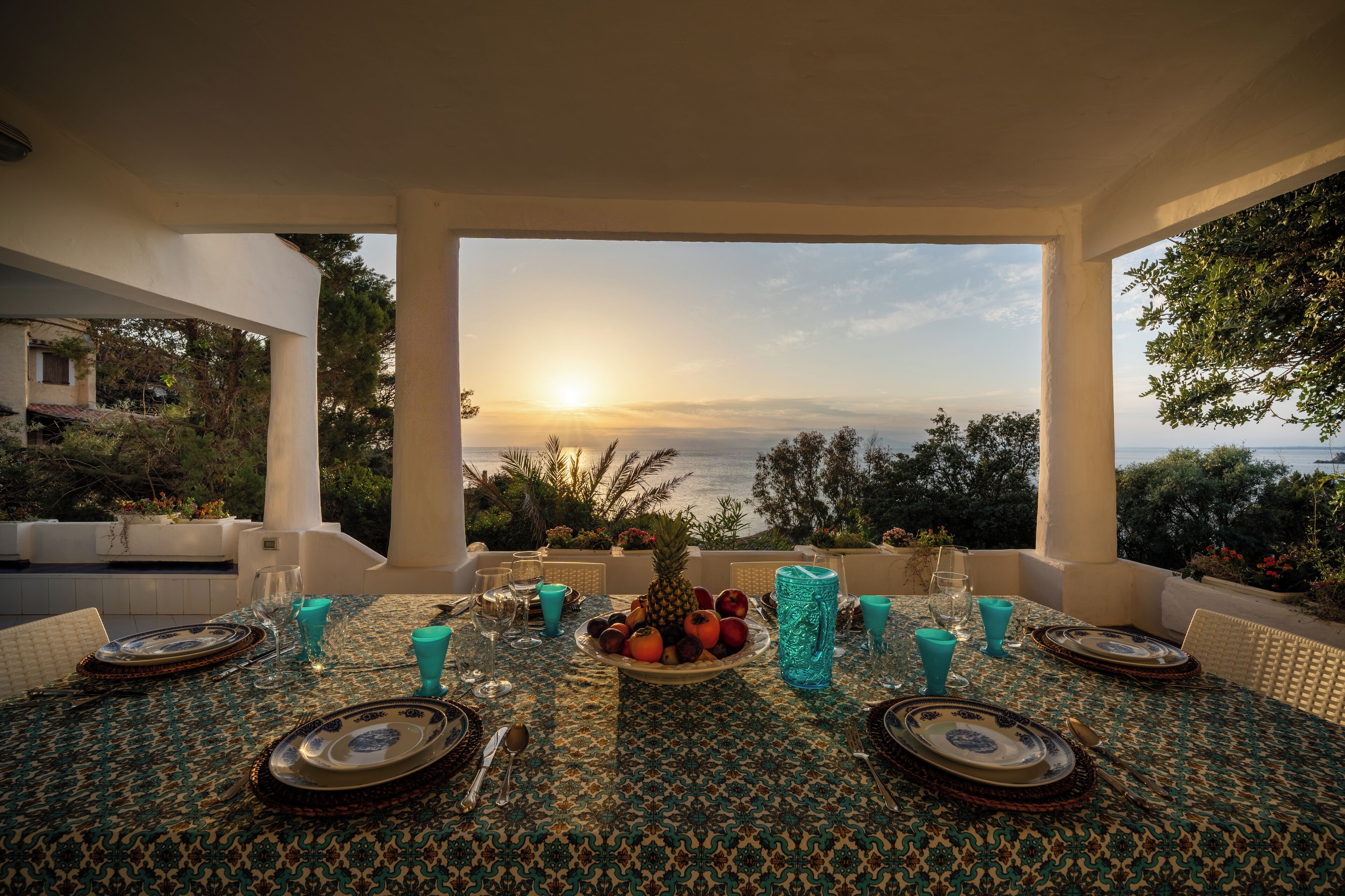 A table set for dining with a view of the sunset over the sea, surrounded by greenery.