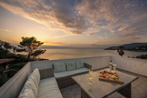 Une terrasse avec un canapé en osier, une table en verre sur laquelle sont posés une bouteille et un plateau de fruits, et une vue sur la mer au coucher du soleil.
