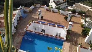 A rooftop pool area with a blue pool, surrounded by white buildings with terracotta roofs, and potted plants.