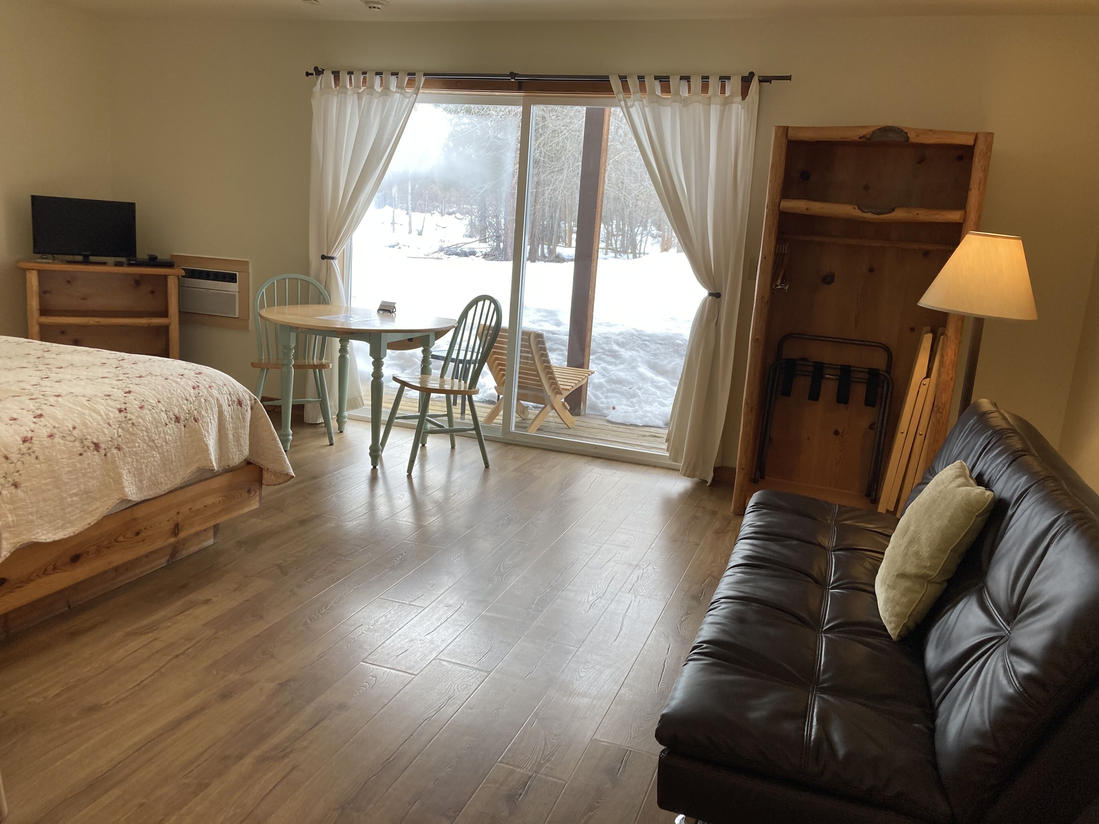 A cozy room with a bed, a leather couch, a table with chairs, a TV, and a window with curtains.
