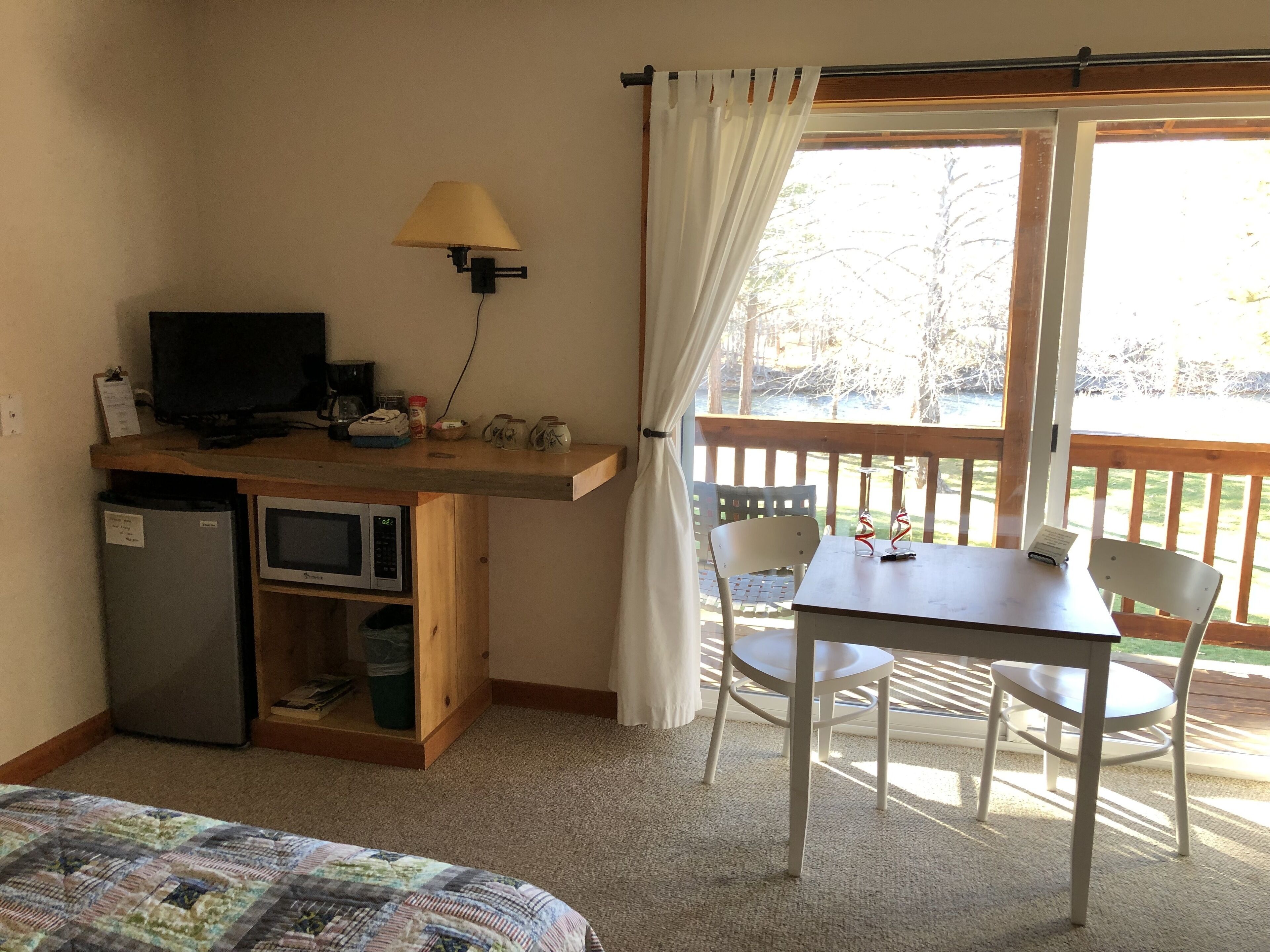 A room with a kitchenette, a television, a table with chairs, and a balcony with a view of trees.