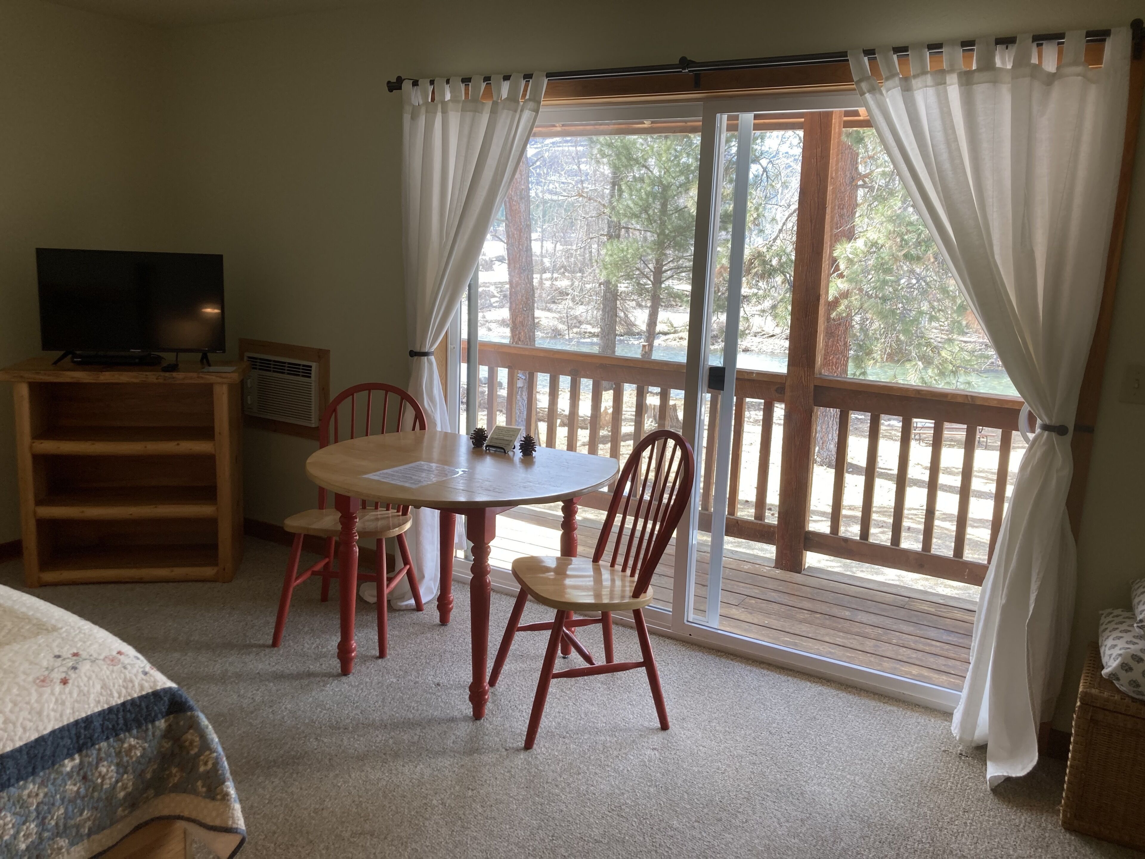 Une pièce avec une table et des chaises en bois, un téléviseur, et une vue sur une terrasse en bois et des arbres, visible à travers de grandes fenêtres.