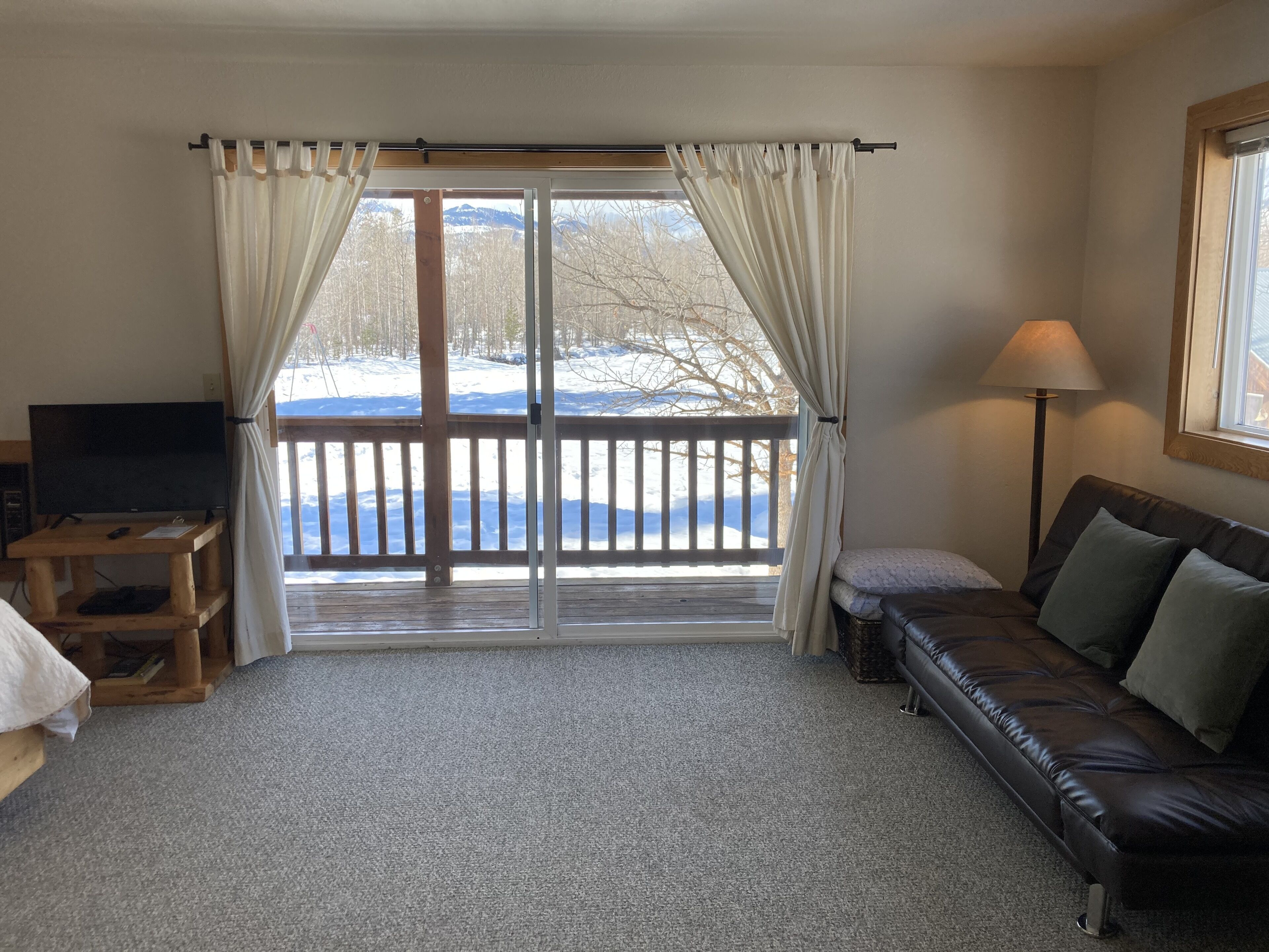 A cozy living room with a large sliding glass door leading to a snowy outdoor area, a leather sofa, a wooden table with a TV, and a lamp.