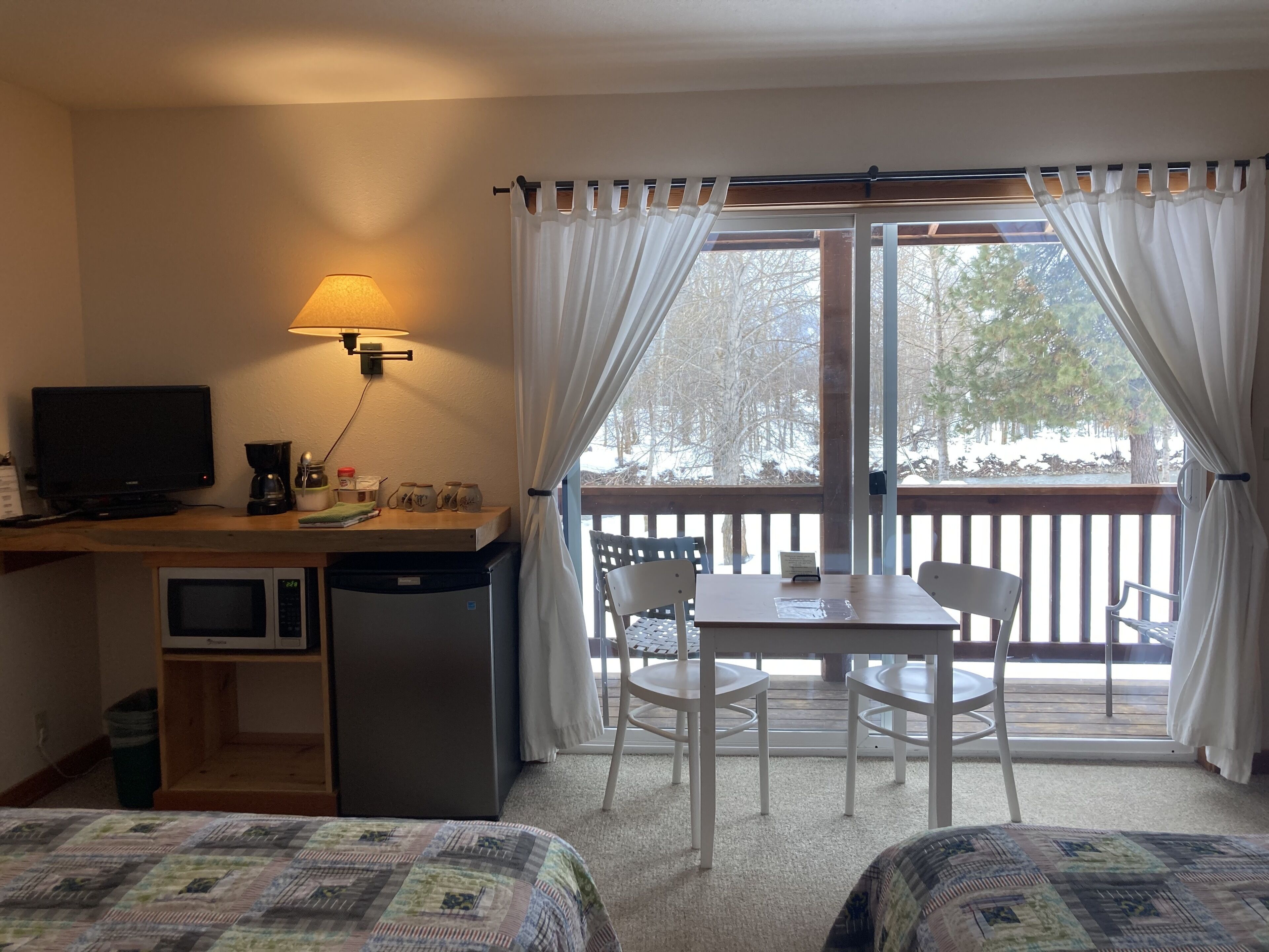 Habitación de hotel con escritorio, televisor, microondas y un pequeño refrigerador. Tiene un balcón con mesa y sillas, y se ve arbolado al exterior.