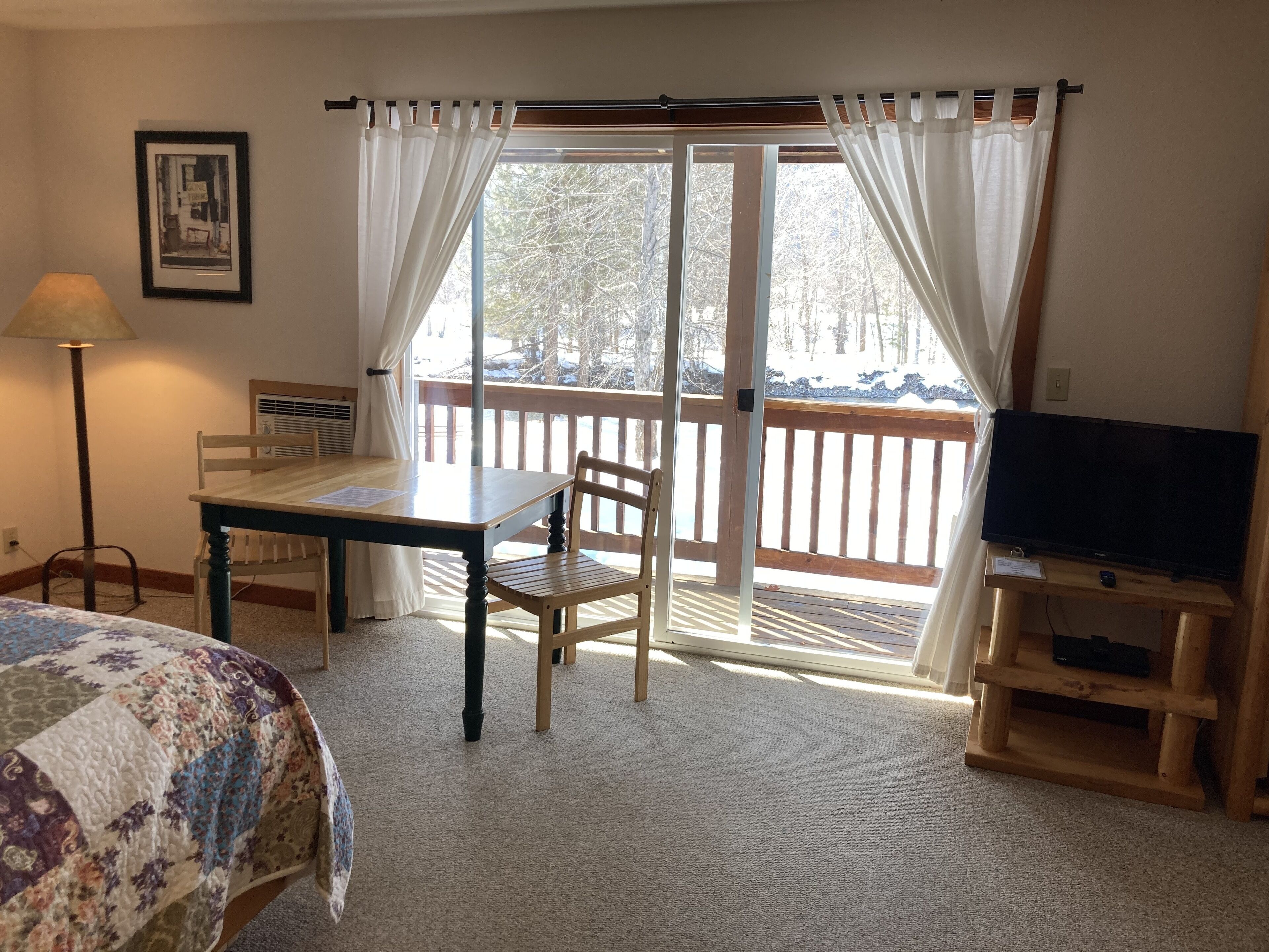 A room with a wooden table, two chairs, a television on a wooden stand, a framed picture on the wall, and a window with curtains.
