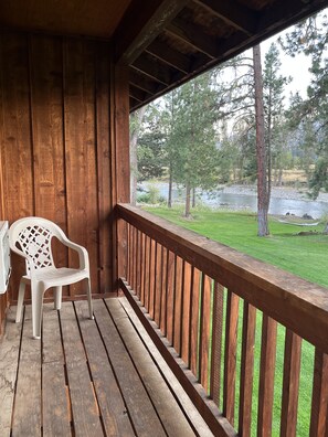 A wooden deck with a white plastic chair and a table, overlooking a grassy area and a body of water with trees in the background.