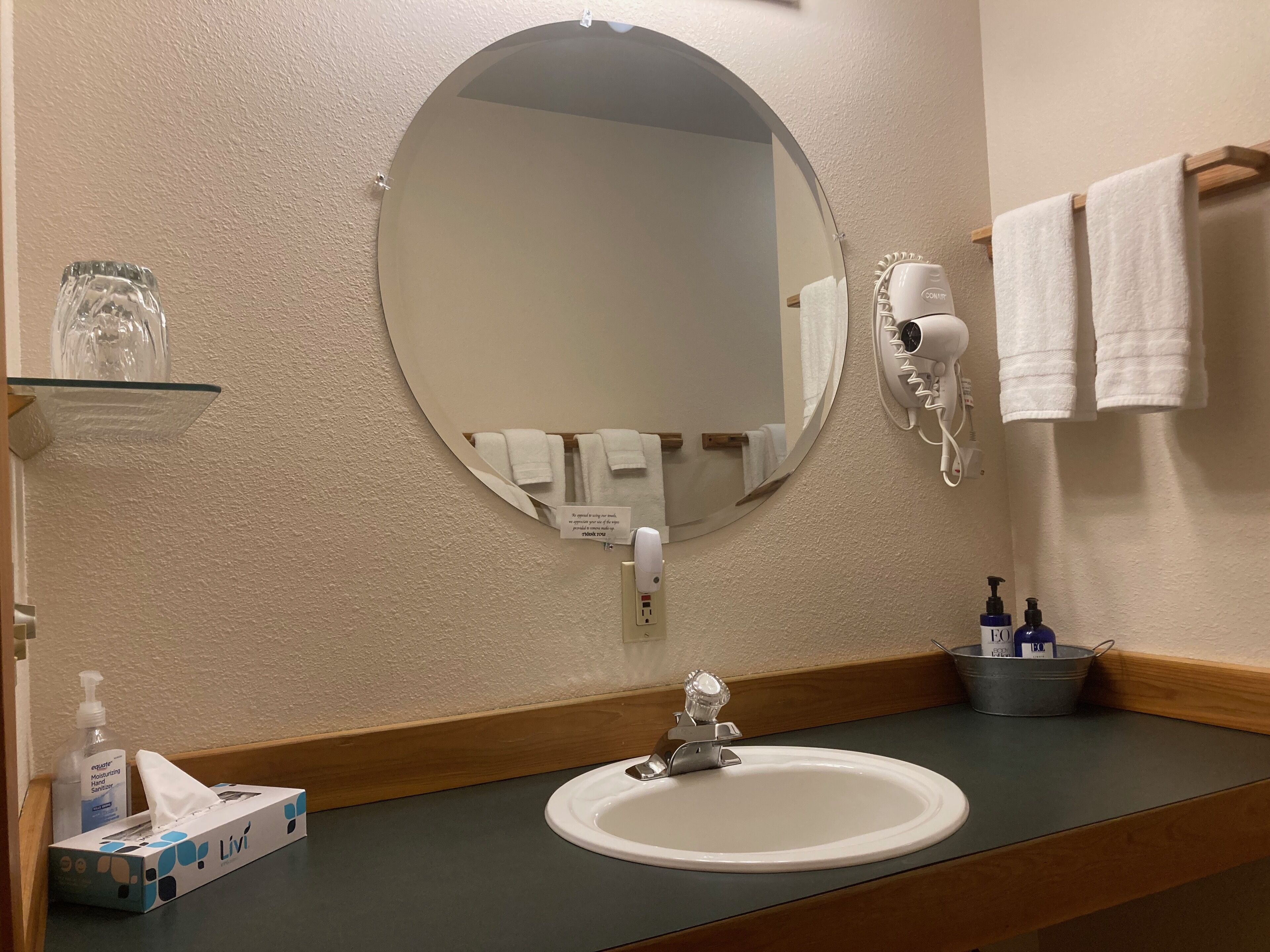 Une salle de bain équipée d’un miroir rond, d’un lavabo, d’un porte-serviettes, d’un sèche-cheveux et d’articles de toilette.