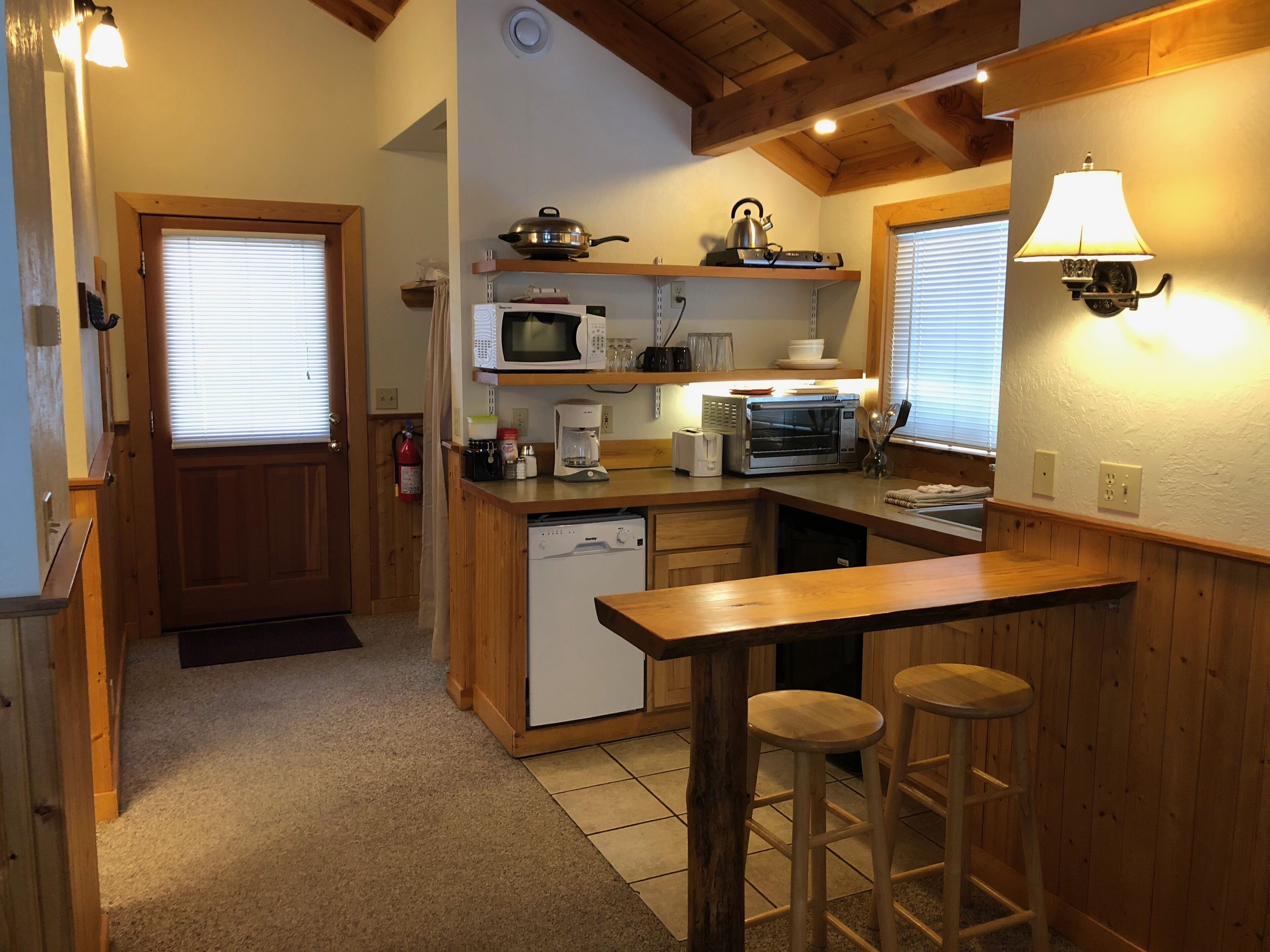 A compact kitchen with wooden countertops, a built-in dishwasher, a microwave, a kettle, and a coffee maker. There are two stools at the bar counter.