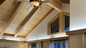 Una habitación acogedora con techo inclinado, ventilador de madera, un amplio ventanal con cortinas, una mesa de madera y un banco acolchado.
