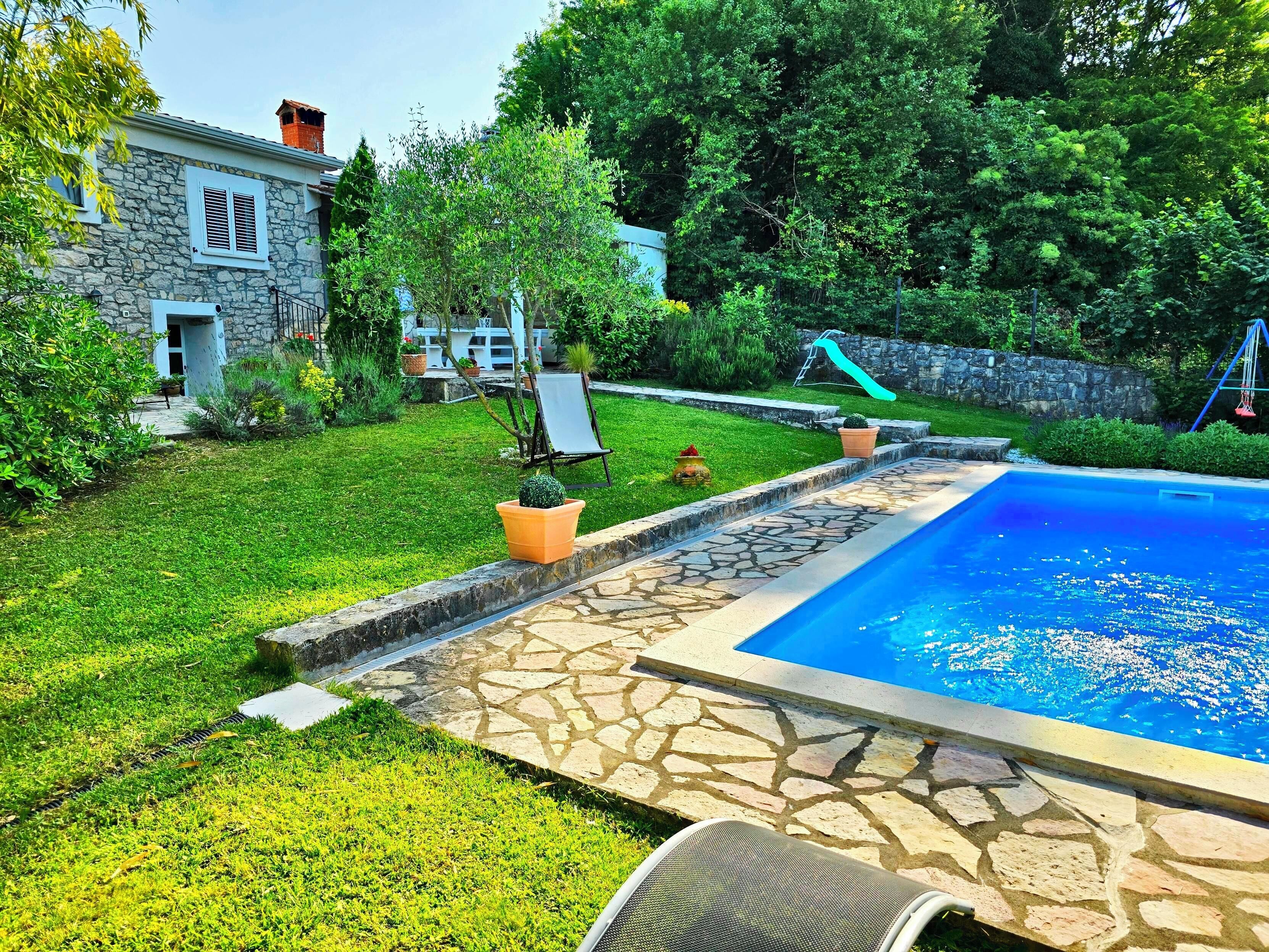 A backyard with a swimming pool, stone patio, and a house with a stone facade.