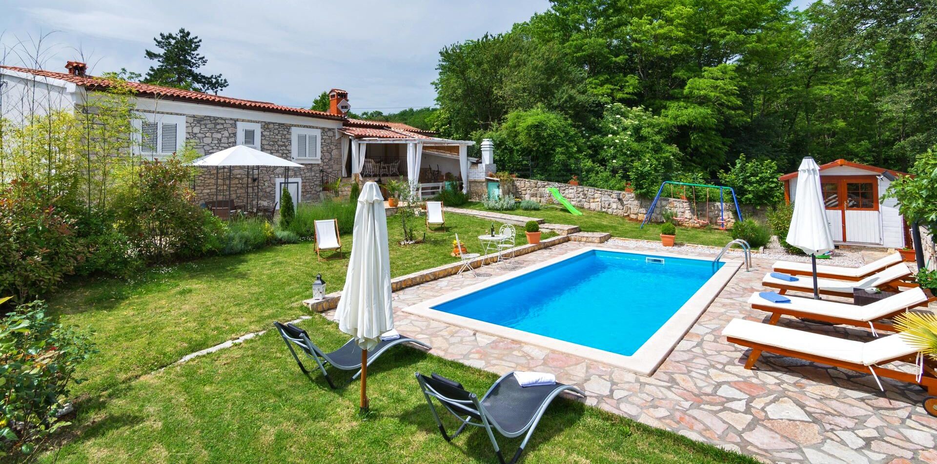 A backyard with a swimming pool, stone patio, lounge chairs, and a covered seating area.