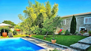 Une piscine aux bords recouverts de pierres, entourée d’une pelouse bien entretenue, d’une maison en pierre et de diverses plantes.