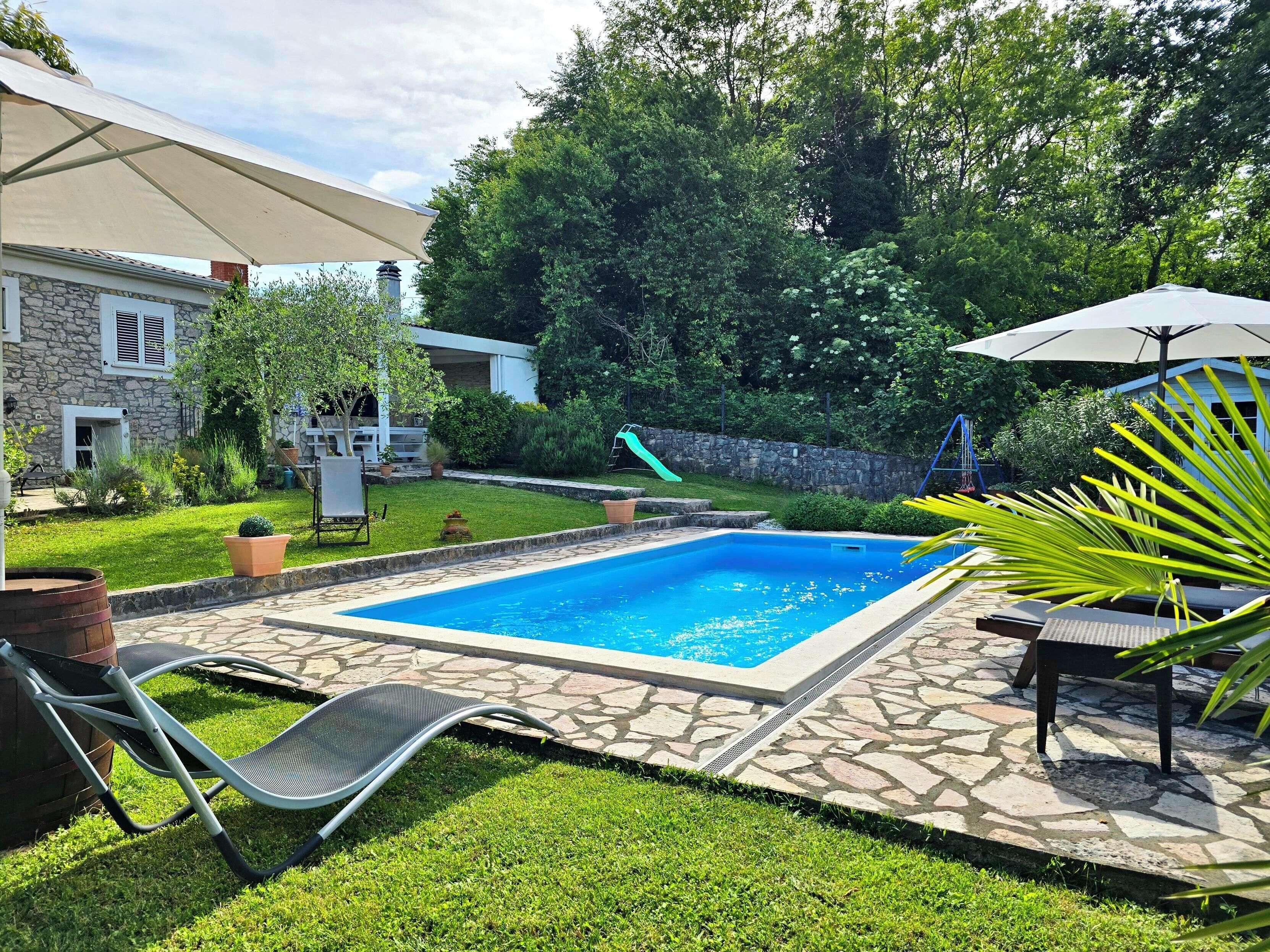Un jardin avec une piscine, une terrasse en pierre, une chaise longue et une table avec un parasol.