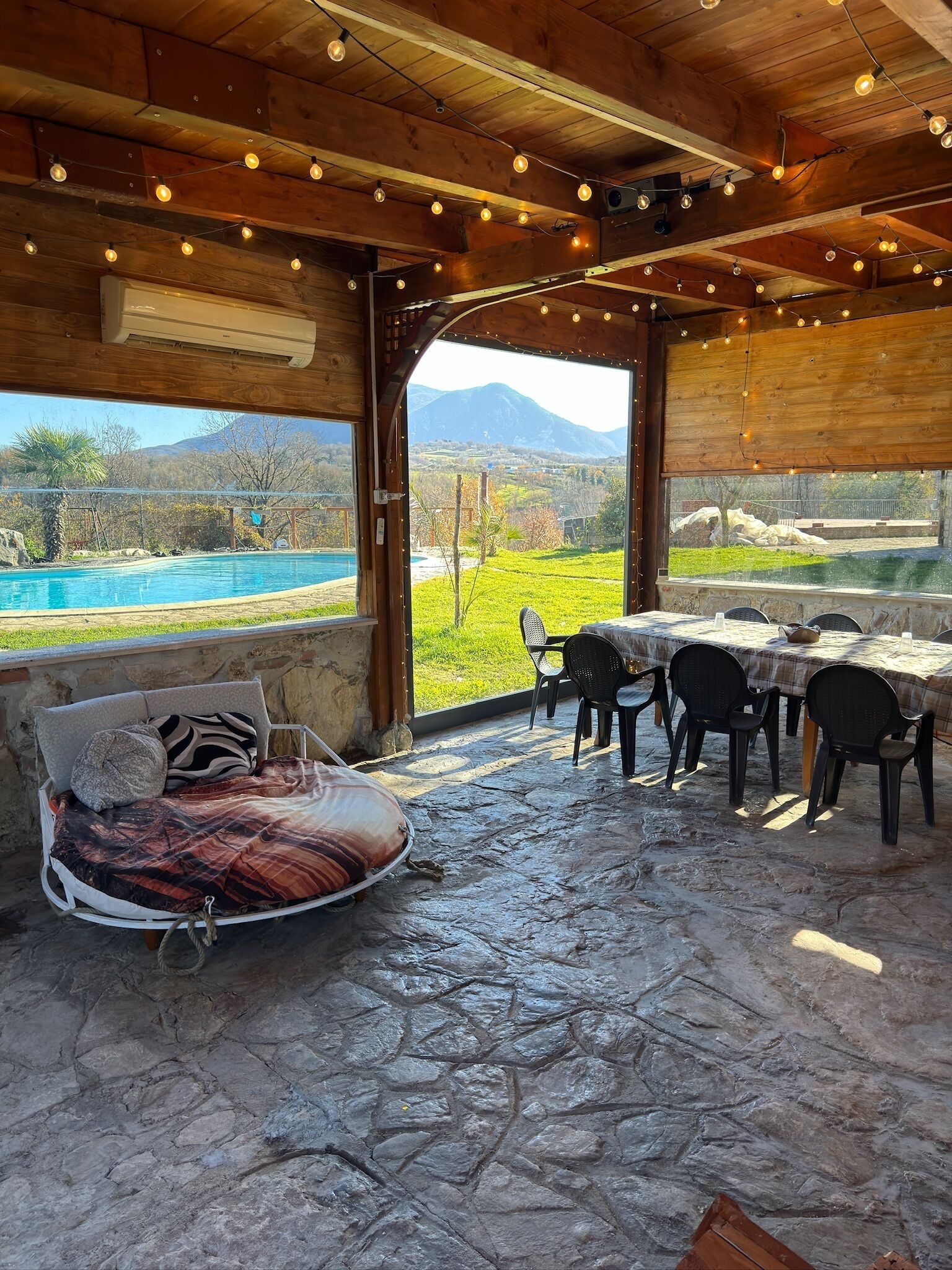 A rustic wooden interior with a stone floor, a bed with a patterned blanket, a dining table with chairs, and a view of a mountain and pool outside.